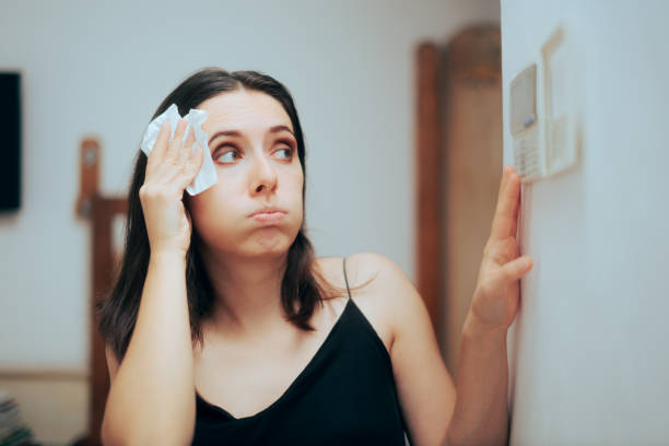 A woman wipes sweat from the side of her head while attempting to change the temperature of the thermostat.